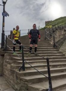 Image of two smiling men, wearing running clothes, standing on the steps up to Whitby Abbey.